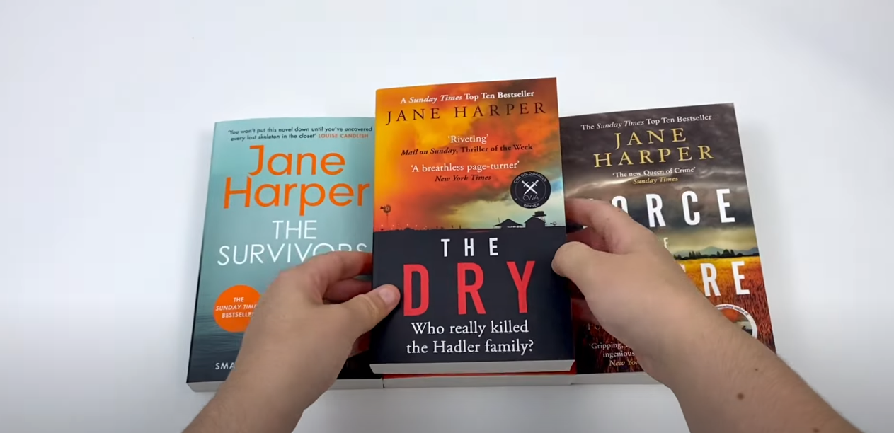 Hand Holding 'The Dry' by Jane Harper, Alongside Other Jane Harper Books on Table in the Background